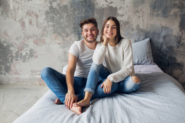 Junges lächelndes Paar, das zu Hause im lässigen Outfit auf Bett sitzt, Mann und Frau, die Spaß zusammen haben, glücklich