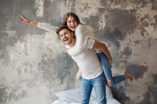 Junges lächelndes Paar, das zu Hause im lässigen Outfit auf Bett spielt, Mann und Frau, die Spaß zusammen haben, verrückte positive Emotion, glücklich, Hand hochhaltend