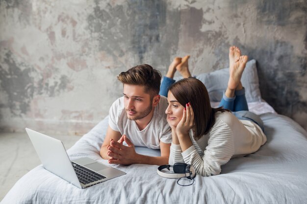 Junges lächelndes Paar, das zu Hause im lässigen Outfit auf dem Bett liegt und in Laptop, Mann und Frau schaut, die glückliche Zeit zusammen verbringen, entspannend
