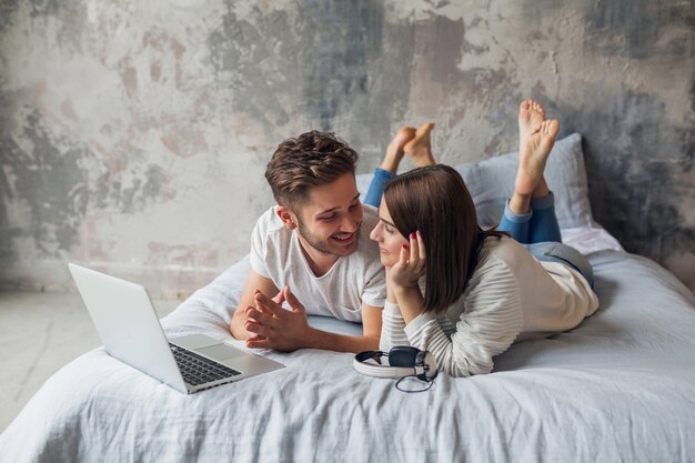Junges lächelndes Paar, das zu Hause im lässigen Outfit auf dem Bett liegt und in Laptop, Mann und Frau schaut, die glückliche Zeit zusammen verbringen, entspannend