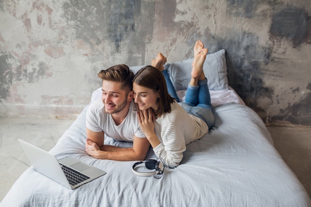 Junges lächelndes Paar, das zu Hause im lässigen Outfit auf dem Bett liegt und in Laptop, Mann und Frau schaut, die glückliche Zeit zusammen verbringen, entspannend