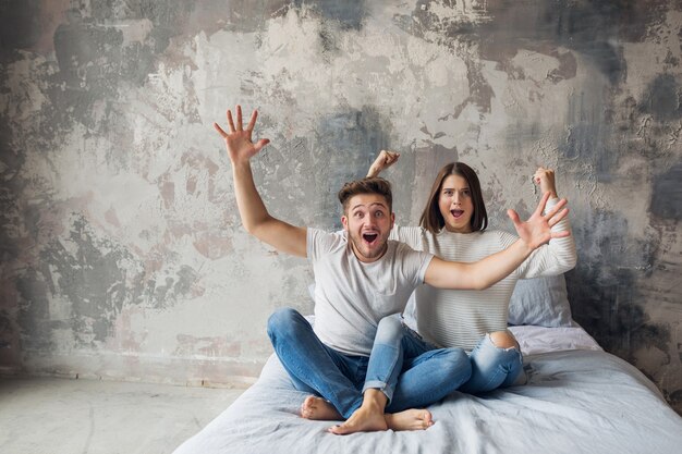 Junges lächelndes Paar, das zu Hause im lässigen Outfit auf dem Bett sitzt, Mann und Frau, die Spaß zusammen haben, verrückte positive Emotion, glücklich, Hände hoch haltend