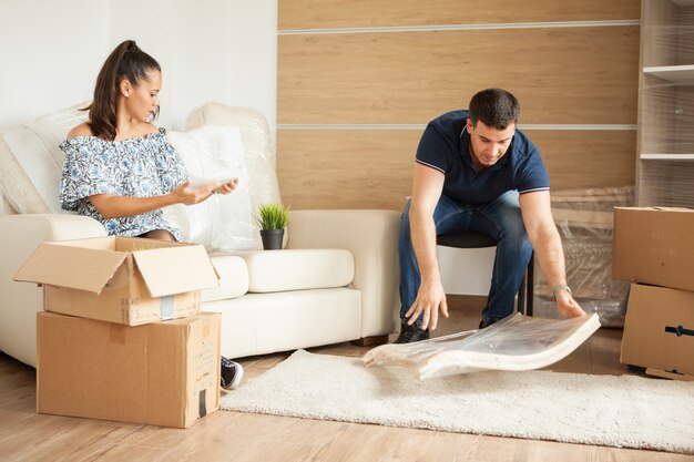 Junges lächelndes Paar, das zusammen auf Sofa sitzende Kartons auspackt. Sitzen im Wohnzimmer.