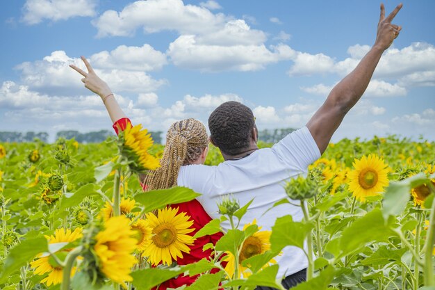 Junges Liebespaar, das mitten in einem Sonnenblumenfeld steht und ein Siegeszeichen zeigt