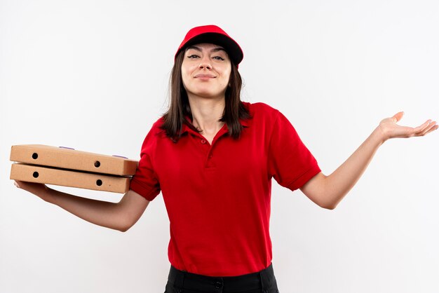 Junges Liefermädchen, das rote Uniform und Kappe hält, die Pizzaschachteln hält, die verwirrten lächelnden sich ausbreitenden Arm zur Seite stehen, die über weißer Wand steht