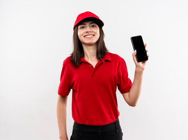 Junges Liefermädchen, das rote Uniform und Kappe trägt, die lächelnd lächelndes Smartphone über weißer Wand zeigt
