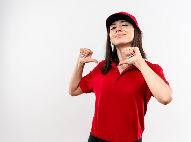 Junges Liefermädchen, das rote Uniform und Kappe trägt, zeigt auf sich selbst lächelnd zuversichtlich stehend über weißer Wand