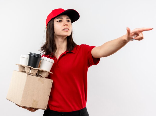 Junges Liefermädchen, das rote Uniform- und Kappenhalteboxpaket und Kaffeetassen trägt, die beiseite schauen auf etwas mit Zeigefinger stehen über weißem Hintergrund