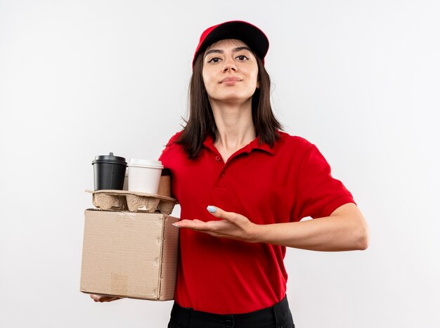 Junges Liefermädchen, das rote Uniform und Kappenhalteboxpaket und Kaffeetassen trägt, die mit Arm der Hand, die sicher steht, über weißer Wand stehen