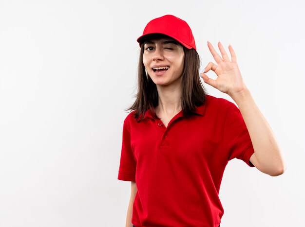 Junges Liefermädchen, das rote Uniform und Mütze Lookign an der Kamera trägt, die mit dem glücklichen Gesicht zwinkert, das ok Zeichen zeigt, das über weißem Hintergrund steht