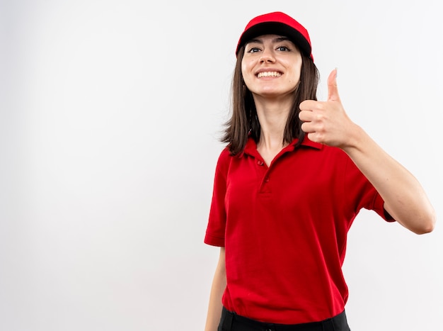 Junges Liefermädchen, das rote Uniform und Mützenlookign an der Kamera trägt, die mit glücklichem Gesicht lächelt, das Daumen oben zeigt über weißem Hintergrund