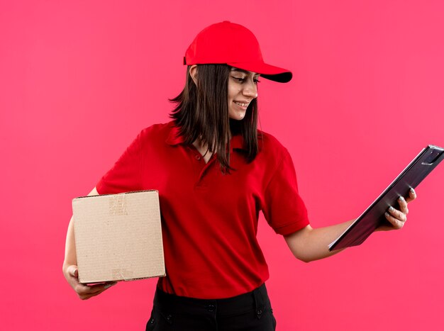 Junges liefermädchen, das rotes poloshirt und kappe hält boxpaket und zwischenablage, die es lächelnd mit glücklichem gesicht über rosa wand stehend betrachten