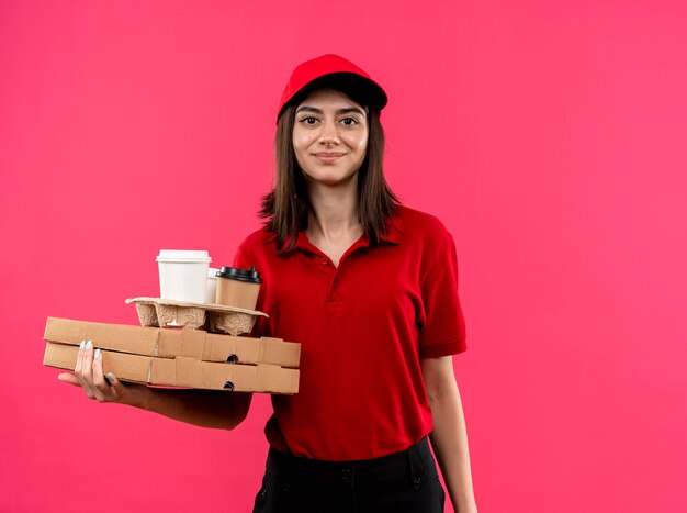 Junges Liefermädchen, das rotes Poloshirt und Kappe hält, die Pizzaschachteln und Nahrungsmittelpaket hält, das mit glücklichem Gesicht steht, das über rosa Wand steht