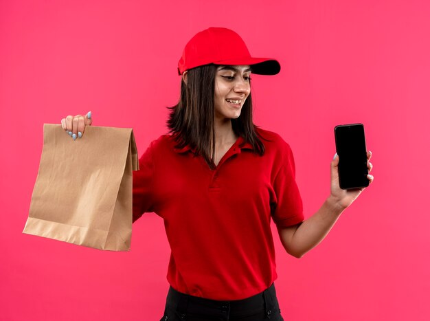Junges Liefermädchen, das rotes Poloshirt und Kappe hält, die Smartphone- und Papierpaket hält, das beiseite mit Lächeln auf Gesicht steht, das über rosa Wand steht