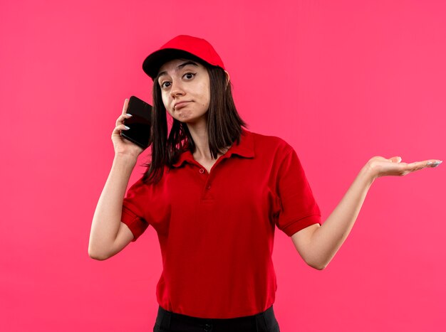 Junges Liefermädchen, das rotes Poloshirt und Kappe trägt, die verwirrten sich ausbreitenden Arm zur Seite schaut, während auf Mobiltelefon steht über rosa Hintergrund