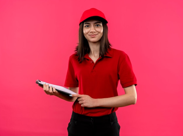 Junges Liefermädchen, das rotes Poloshirt und lächelnde Mütze hält, die über rosa Wand stehend hält