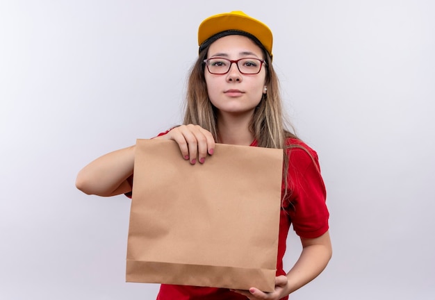 Junges Liefermädchen im roten Poloshirt und in der gelben Kappe, die Papierpaket hält Kamera mit ernstem selbstbewusstem Ausdruck hält