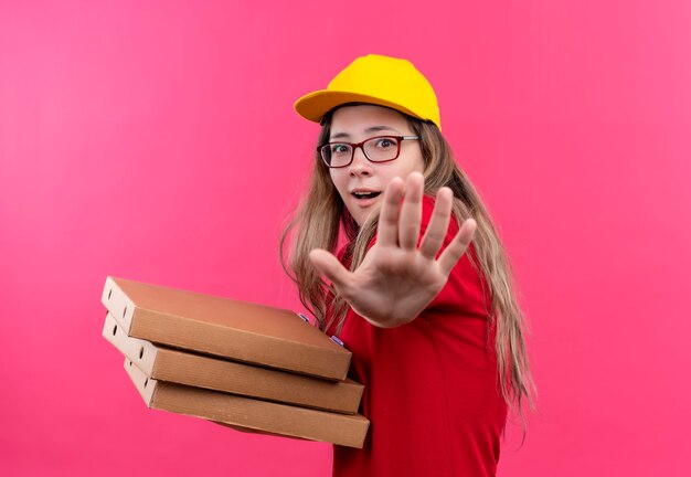 Junges Liefermädchen im roten Poloshirt und in der gelben Kappe, die Stapel von Pizzaschachteln hält, die Verteidigungsgeste mit offener Handfläche machen, ängstlich