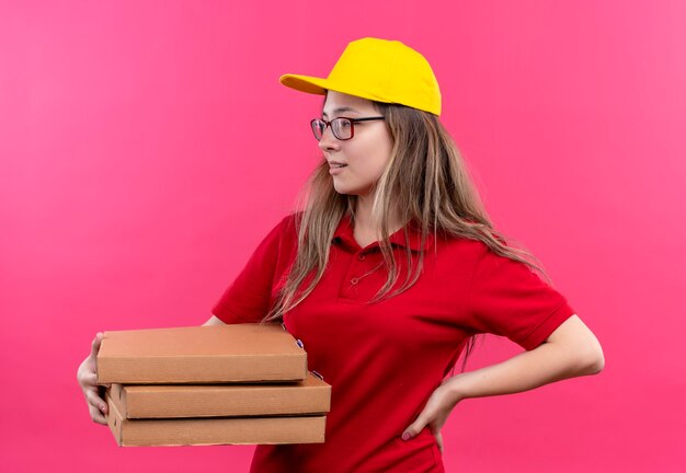 Junges Liefermädchen im roten Poloshirt und in der gelben Kappe, die Stapel von Pizzaschachteln halten, die mit sicherem Ausdruck beiseite schauen