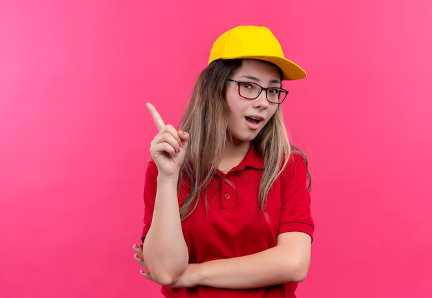 Junges Liefermädchen im roten Poloshirt und in der gelben Kappe, die Zeigefinger lächelnd mit großer neuer Idee zeigt