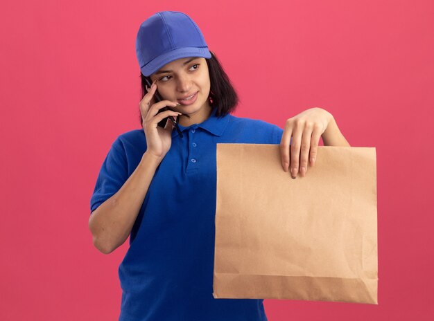 Junges Liefermädchen in blauer Uniform und Kappe, die Papierpaket hält, das auf Handy mit Lächeln auf Gesicht steht, das über rosa Wand steht