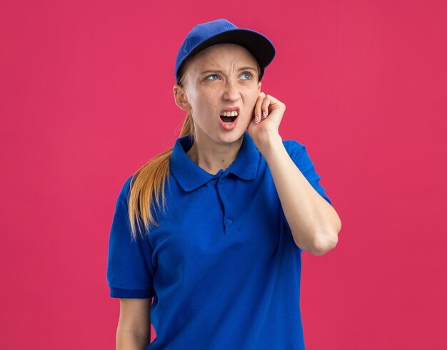 Junges Liefermädchen in blauer Uniform und Mütze, das verwirrt und unzufrieden aufschaut