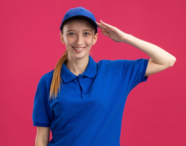 Junges Liefermädchen in blauer Uniform und Mütze lächelnd selbstbewusst salutierend über rosa Wand stehend