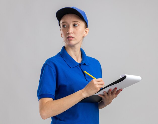 Junges Liefermädchen in blauer Uniform und Mütze mit Bleistift und Klemmbrett mit leeren Seiten, die mit ernstem Gesicht beiseite schauen und etwas über weiße Wand schreiben