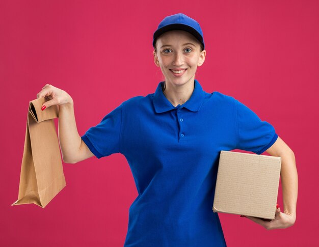 Junges Liefermädchen in blauer Uniform und Mütze mit Karton und Papierpaket, das selbstbewusst über rosa Wand steht