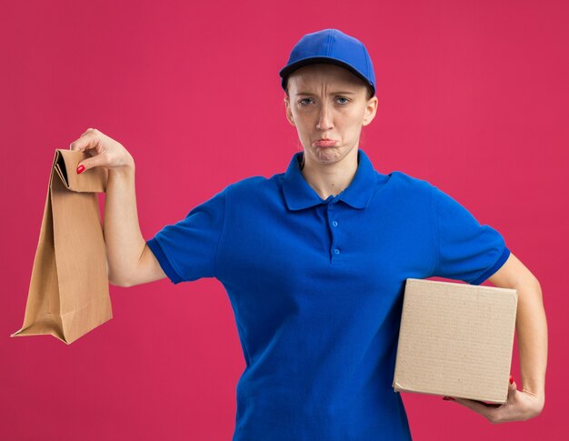 Junges Liefermädchen in blauer Uniform und Mütze mit Pappkarton und Papierpaket mit traurigen Lippen, die über rosa Wand stehen