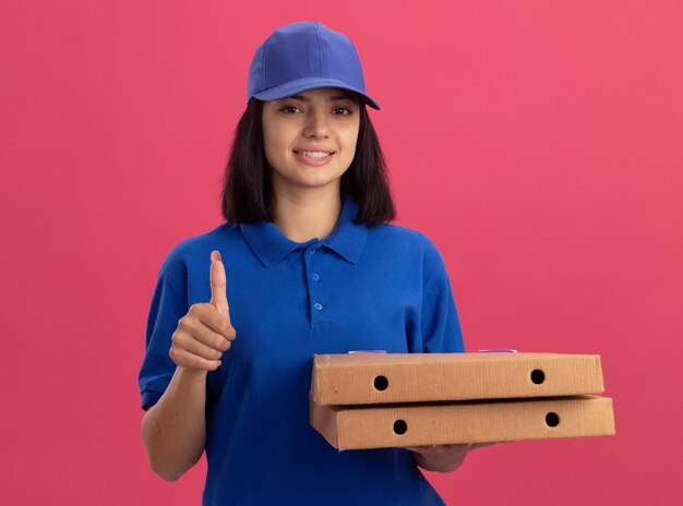 Junges Liefermädchen in der blauen Uniform, die Pizzaschachteln hält, die Daumen oben lächelnd freundlich stehend über rosa Wand zeigen