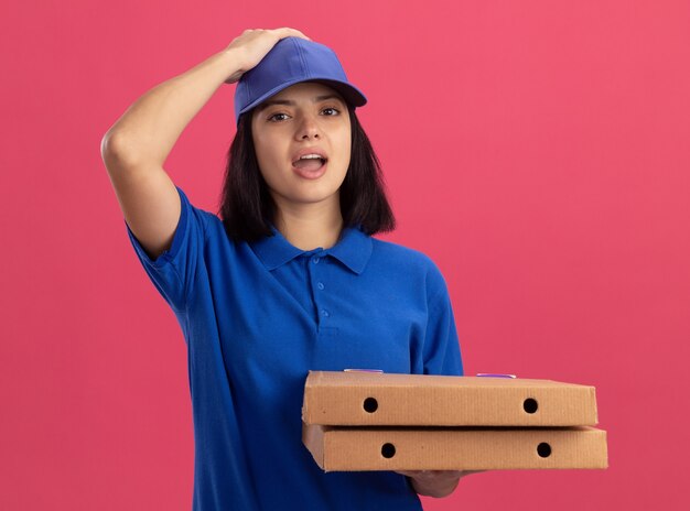 Junges Liefermädchen in der blauen Uniform, die Pizzaschachteln hält, die mit der Hand auf ihrem Hed für den Fehler verwirrt stehen, der über rosa Wand steht