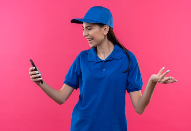 Junges Liefermädchen in der blauen Uniform und in der Kappe, die Bildschirm des Smartphones enttäuscht betrachten