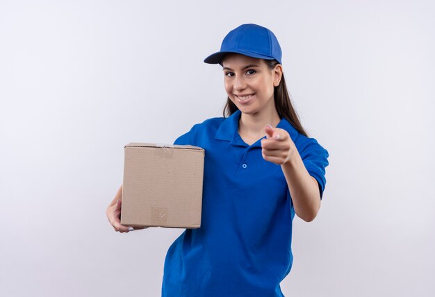 Junges Liefermädchen in der blauen Uniform und in der Kappe, die Box-Paket lächelnd sicheres Zeigen mit dem Finger zur Kamera hält