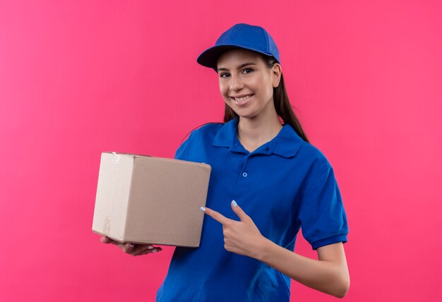 Junges Liefermädchen in der blauen Uniform und in der Kappe, die Boxpaket hält, zeigt mit dem Finger fröhlich samiling