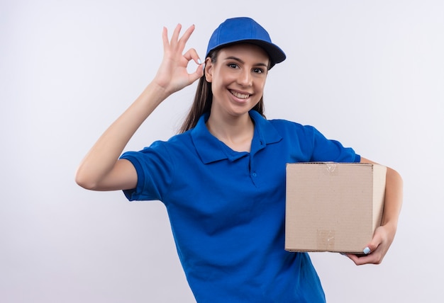 Junges Liefermädchen in der blauen Uniform und in der Kappe, die Boxpaket lächelnd zuversichtlich zeigt, ok Zeichen zeigend