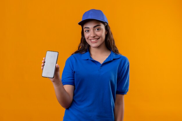 Junges Liefermädchen in der blauen Uniform und in der Kappe, die das lächelnd lächelnde Smartphone über gelbem Hintergrund zeigt