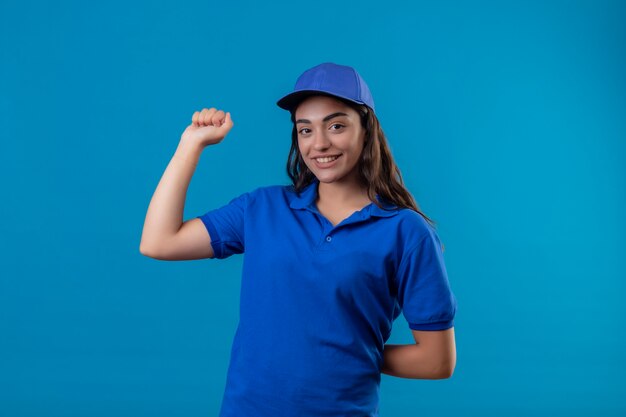 Junges Liefermädchen in der blauen Uniform und in der Kappe, die die Faust anhebt, die Kamera betrachtet, die zuversichtlich lächelt und sich über ihren Erfolg und Sieg freut, die über blauem Hintergrund stehen