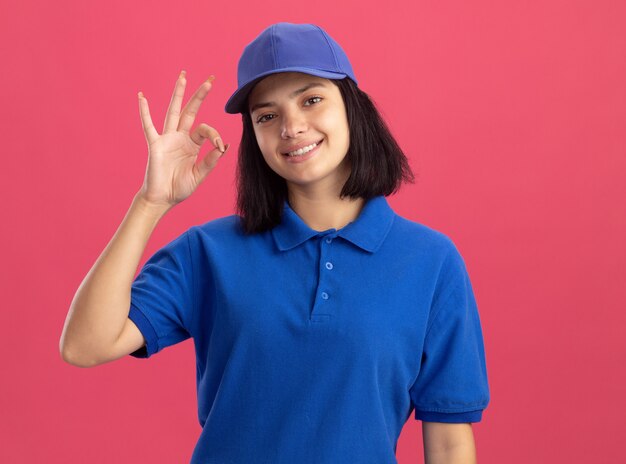 Junges Liefermädchen in der blauen Uniform und in der Kappe, die freundlich lächeln, zeigt okes Zeichen, das über rosa Wand steht