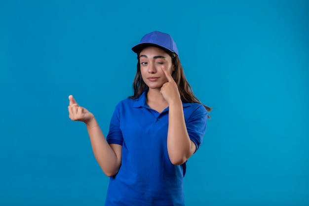 Junges Liefermädchen in der blauen Uniform und in der Kappe, die Geldgestik macht, die zuversichtlich schaut, die Kamera zeigt, zeigt Finger zu ihrem Auge und wartet auf Zahlung, die über blauem Hintergrund steht