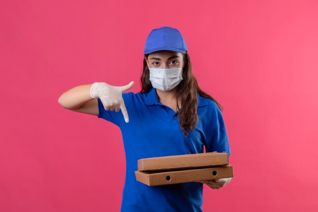 Junges Liefermädchen in der blauen Uniform und in der Kappe, die Gesichtsschutzmaske hält, die Pizzaschachteln hält, die mit Finger auf sie zeigen Kamera mit zuversichtlichem ernstem Ausdruck stehen, der über Stift steht