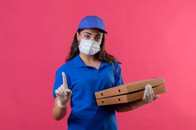 Junges Liefermädchen in der blauen Uniform und in der Kappe, die Gesichtsschutzmaske und Handschuhe hält, die Pizzakästen halten, die Finger oben warnende Warngeste betrachten Kamera mit ernstem Gesicht stehen über pi stehen