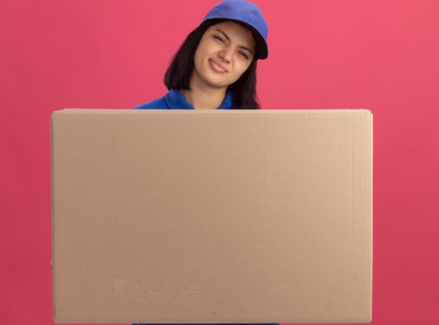 Junges Liefermädchen in der blauen Uniform und in der Kappe, die großen Pappkarton hält, der ironischen Mund mit enttäuschtem Ausdruck macht, der über rosa Wand steht