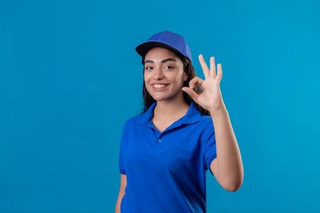 Junges Liefermädchen in der blauen Uniform und in der Kappe, die Kamera betrachten, die fröhlich tut, ok Zeichen, das über blauem Hintergrund steht