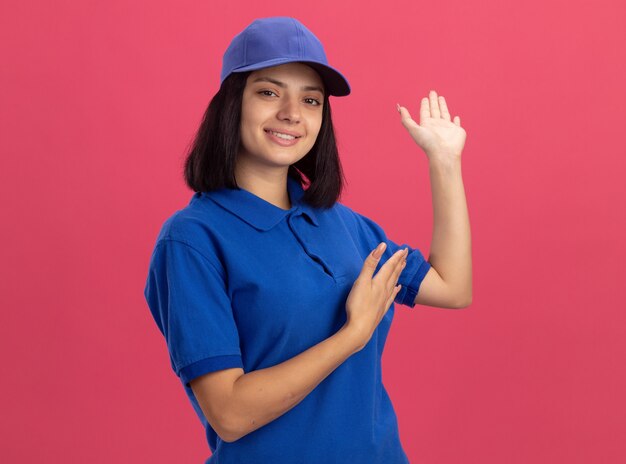 Junges Liefermädchen in der blauen Uniform und in der Kappe, die mit den Armen oh Hände präsentiert, die fröhlich über rosa Wand stehend lächeln