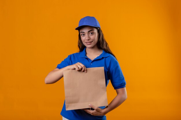 Junges Liefermädchen in der blauen Uniform und in der Kappe, die Papierpaket hält, das freundliches Stehen der Kamera über gelbem Hintergrund betrachtet