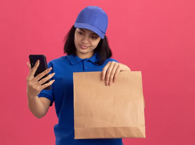 Junges Liefermädchen in der blauen Uniform und in der Kappe, die Papierpaket hält, das ihr Smartphone mit skeptischem Ausdruck betrachtet, der über rosa Wand steht