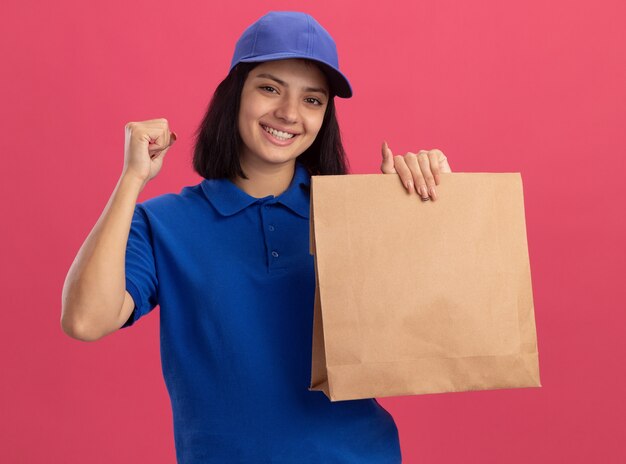 Junges Liefermädchen in der blauen Uniform und in der Kappe, die Papierpaket hält, die Faust glücklich und aufgeregt über rosa Wand stehend
