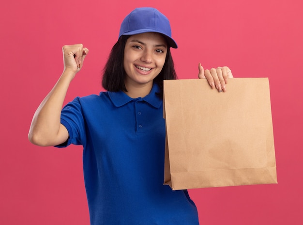 Junges Liefermädchen in der blauen Uniform und in der Kappe, die Papierpaket hält, die Faust glücklich und aufgeregt über rosa Wand stehend