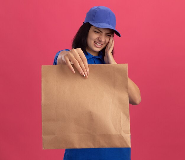 Junges Liefermädchen in der blauen Uniform und in der Kappe, die Papierpaket zeigt, das unwill berührt, das ihren Kopf berührt, der unter Kopfschmerzen steht, die über rosa Wand stehen